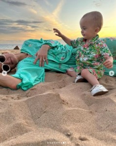 砂浜に寝転ぶパリスのそばに座るフェニックス君。母親を見つめながら、愛らしい笑顔を見せている（『Paris Hilton　Instagram「Magical Memories at ＠GrandWailea that I will cherish forever」』より）